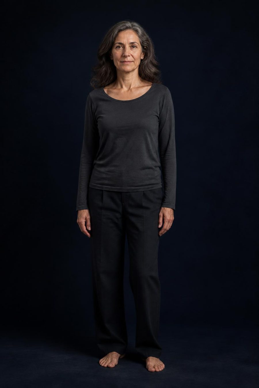 A woman in her early 50s standing centered, full-length, against a deep ink backdrop — calm, present, the whole person.