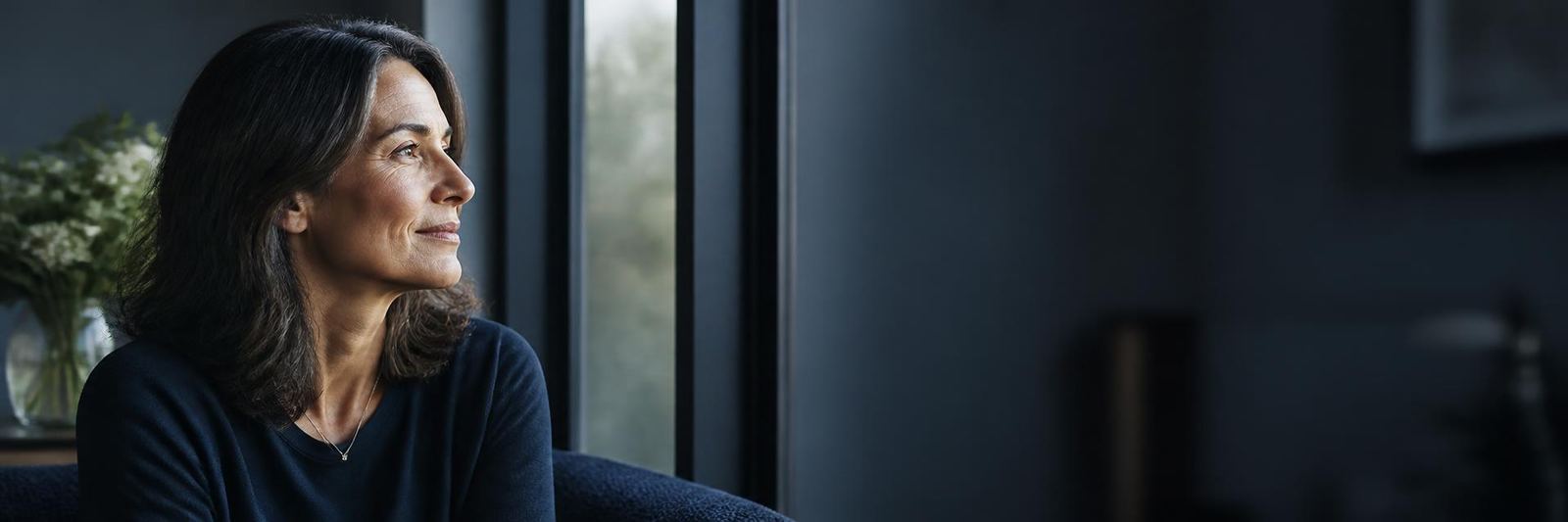 A woman in her early 50s near a window in soft directional light — quiet trust, presence, unhurried.