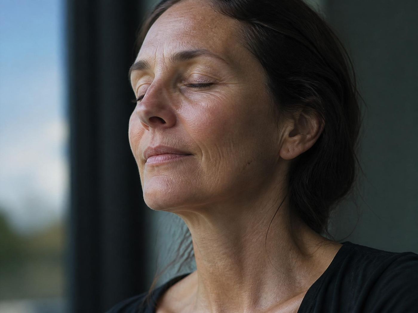 A close-up of a woman in her early 50s in soft window light — real, unretouched skin texture.